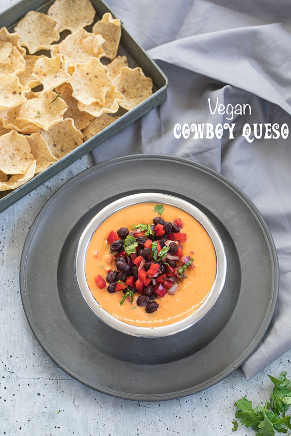 Top view of a stainless steel bowl filled with cowboy queso placed on a gray dish. There is a gray tray next to it filled with tostios scoop chips