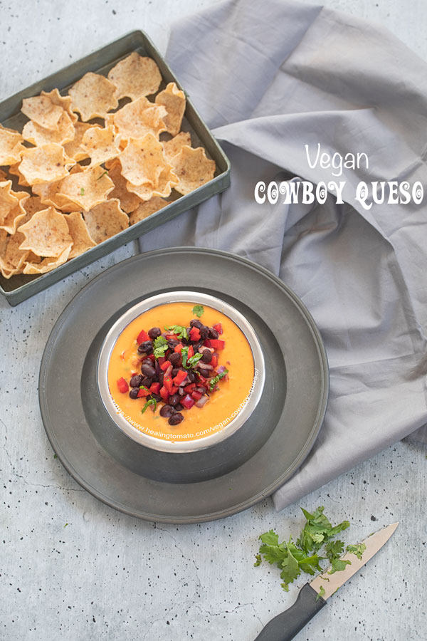 Top view of a stainless steel bowl filled with cowboy queso placed on a gray dish. There is a gray tray next to it filled with tostios scoop chips