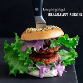 Front view of the double decker everything bagel brekafast burger on a light black stone tray with a knife handle sticking out of the top of the bagel