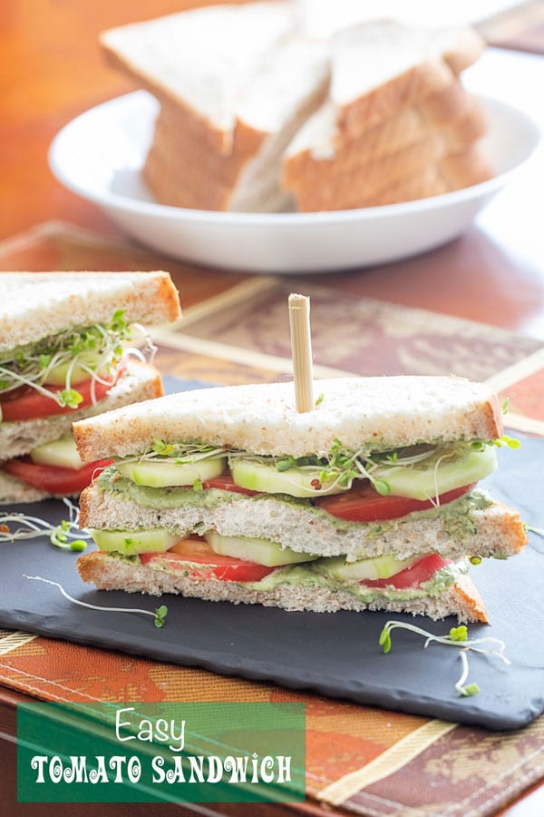 Front view of a double decker tomato sandwich with layers of tomato, cucumber, pesto spread and broccoli sprouts. A bowl of sliced bread is visible in the background