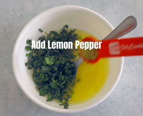 top view of a red measuring spoon adding lemon pepper to a white bowl with butter + herbs