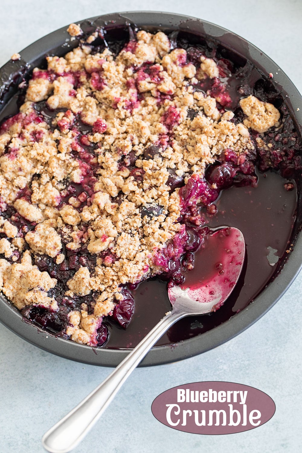 Top view of a partial blueberry crumble pie in a steel pan with a spoon in it.