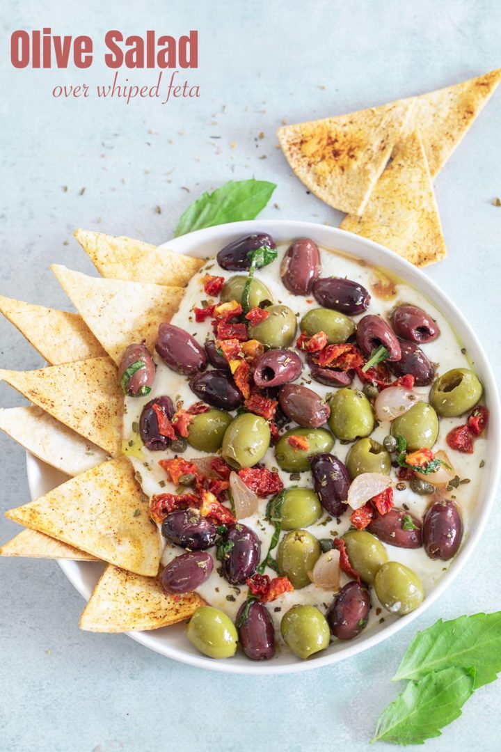 Top, closeup view of olive salad with sun-dried tomatoes on a whipped feta dip with a tortilla chips on inserted on the left side of the dip
