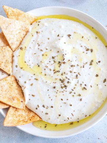 Closeup view of a white bowl filled with the whipped feta bowl with oil and seasoning as garnish. The left side of the plate is decorated with tortilla chips