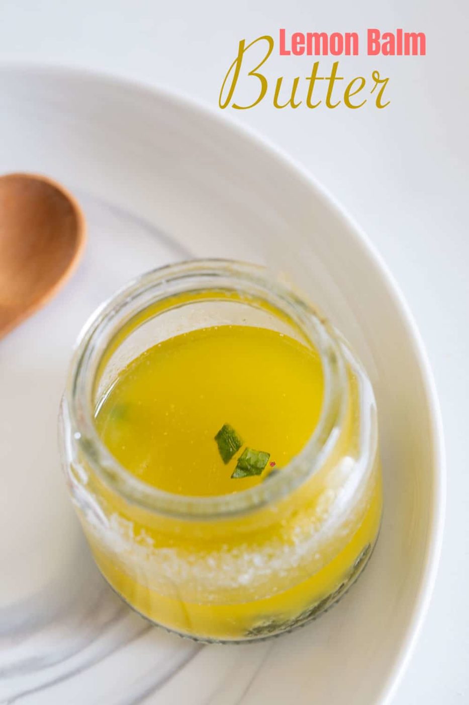 Top view of a glass bottle filled with melted lemon balm butter and few chopped lemon balm pieces floating on top. A wooden spoon next to the bottle.