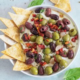 Top, closeup view of olive salad with sun-dried tomatoes on a whipped feta dip with a tortilla chips on inserted on the left side of the dip