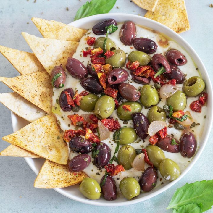 Top, closeup view of olive salad with sun-dried tomatoes on a whipped feta dip with a tortilla chips on inserted on the left side of the dip