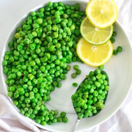 Closeup vew of a white plate filled with cooked green peas and a serving spoon with lemon rounds on top of the plate.
