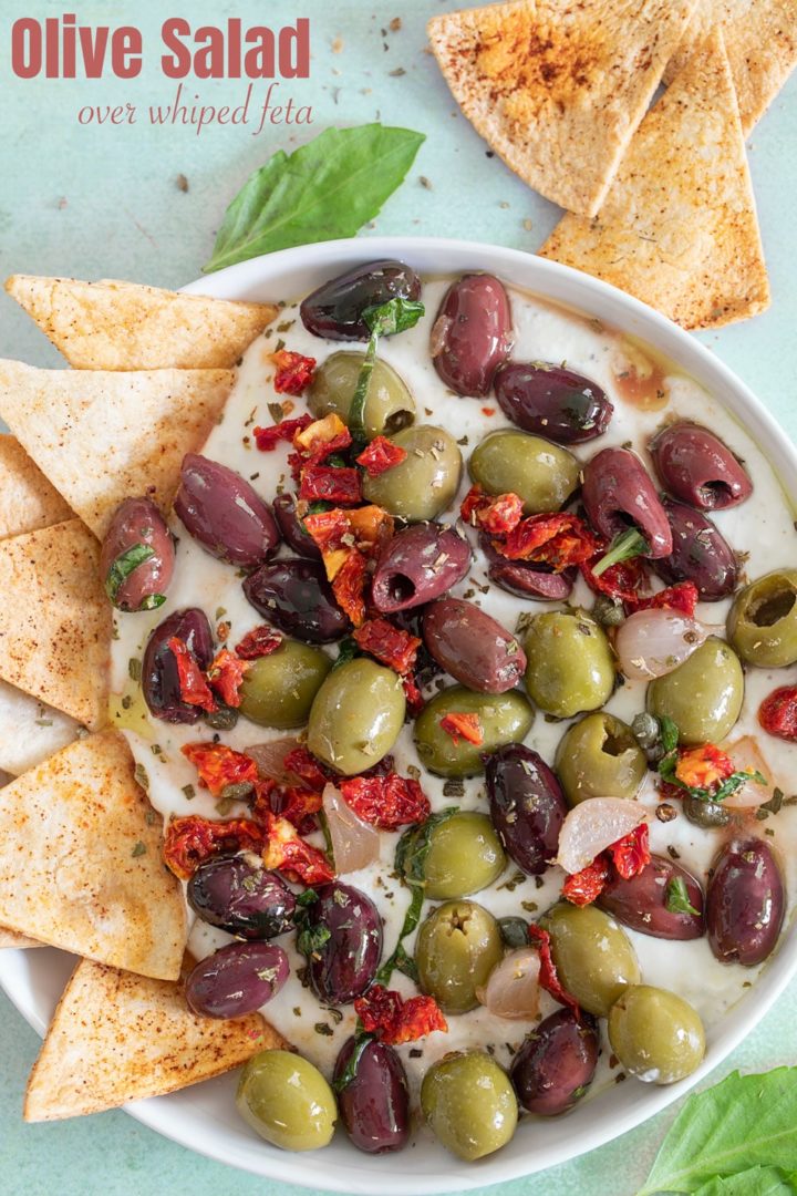 Top view of olive salad on a whipped feta dip with a tortilla chips on inserted on the left side of the dip