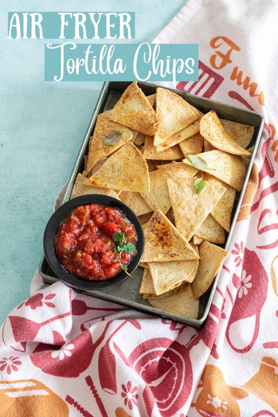 top view of a grey pan filled with air fryer tortilla chips and a salsa plate placed on the inner left side of pan. Grey pan is on top of a colorful kitchen towel