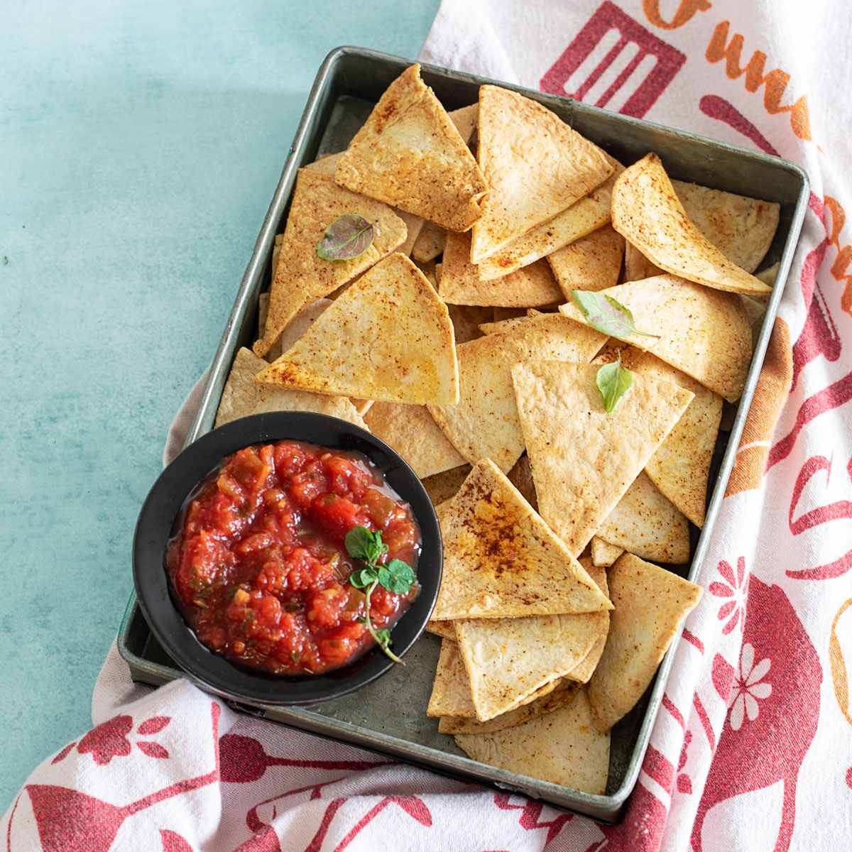 top view of a grey pan filled with air fryer tortilla chips and a salsa plate placed on the inner left side of pan. Grey pan is on top of a colorful kitchen towel
