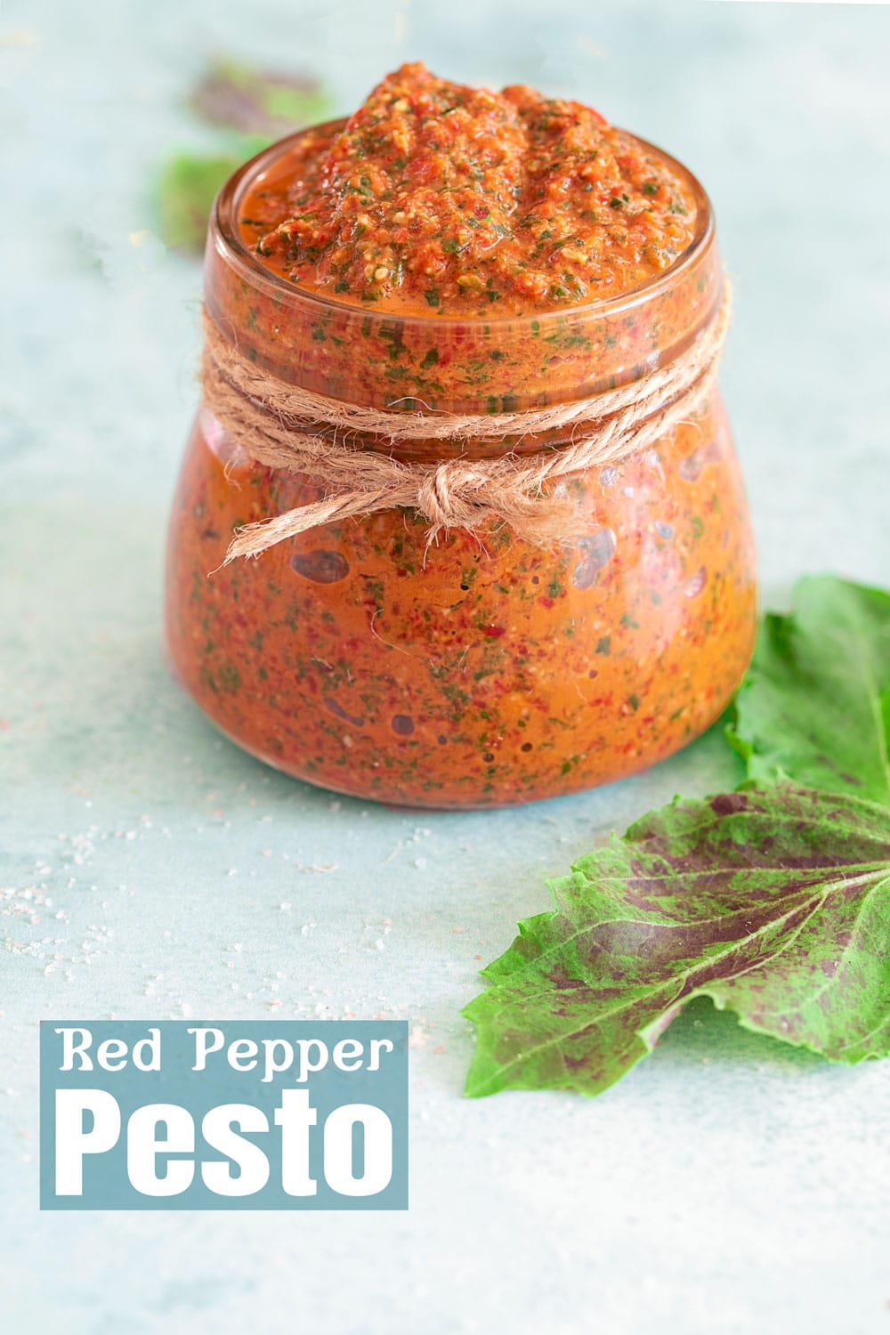 Front view of a small glass jar with a rope tied around the neck of the bottle, filled with pesto. Purplish basil on the right