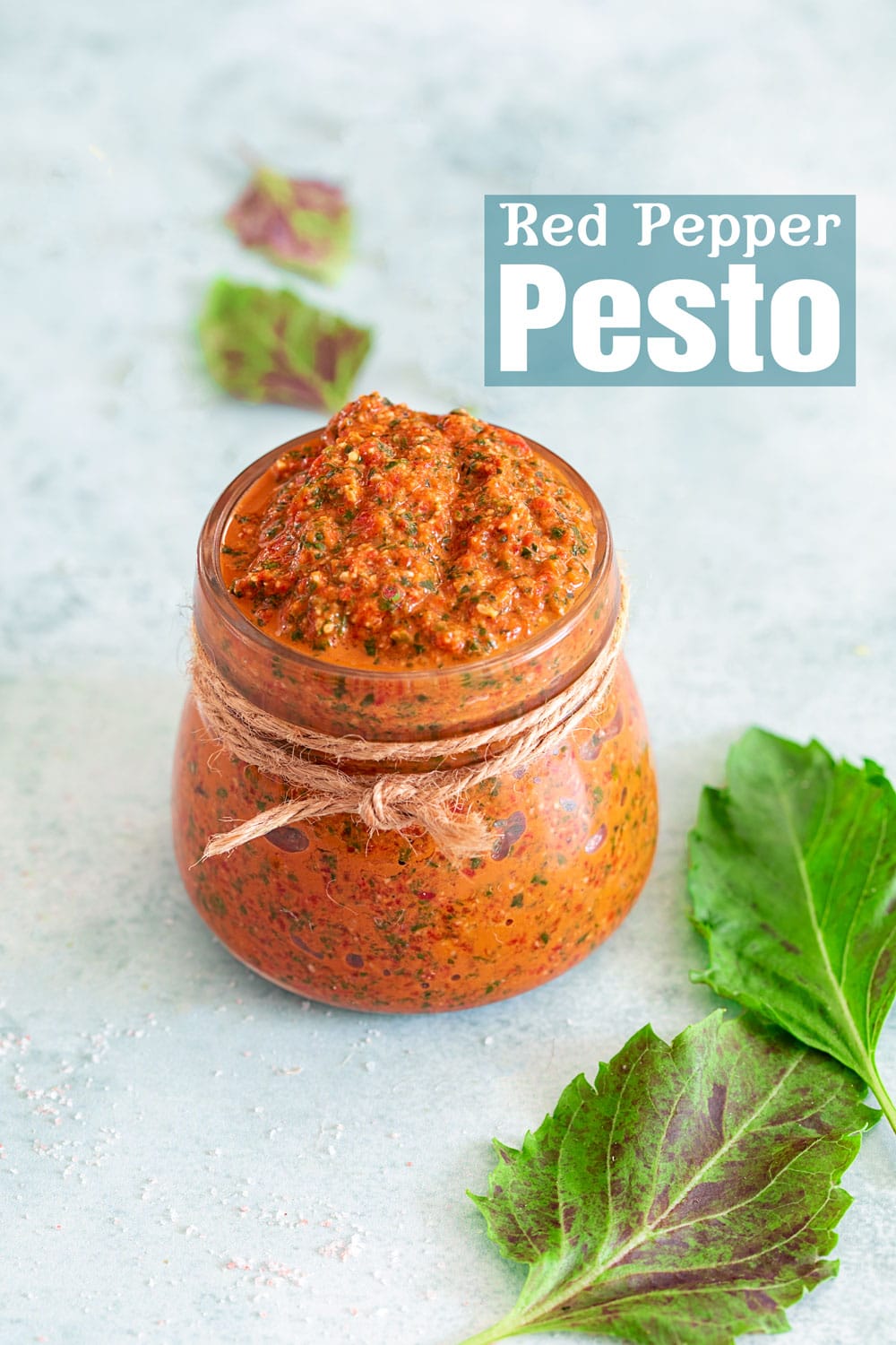 Front view of a small glass jar with a rope tied around the neck of the bottle. Filled with bell pepper pesto.  Purplish basil on the right and in the background