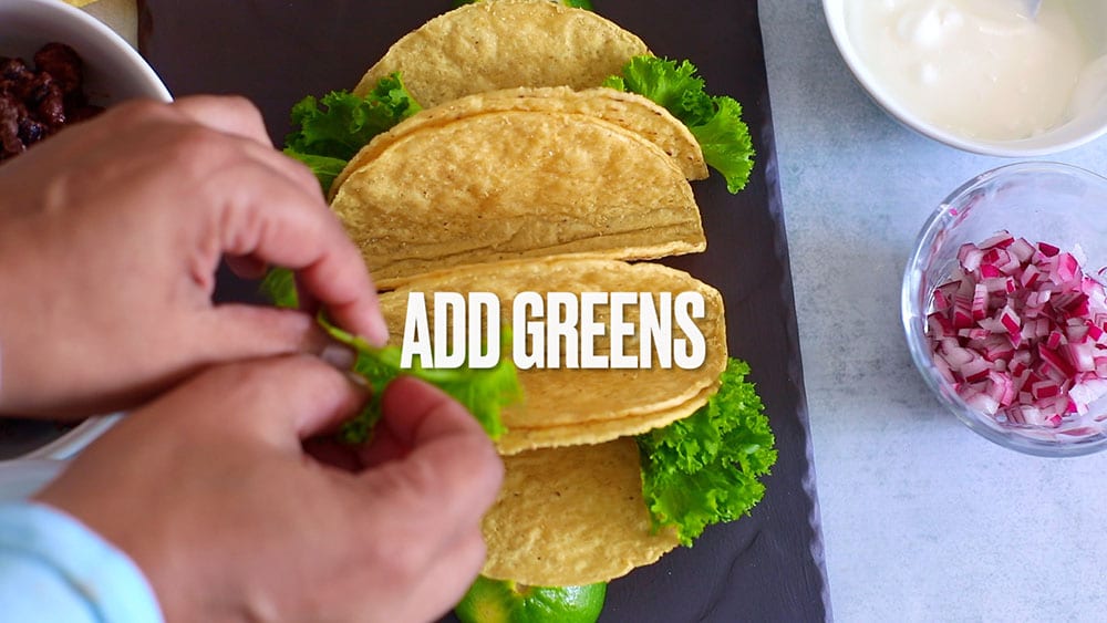 Top view of the author putting greens into 4 taco shells. Bowls containing other ingredients can be seen.