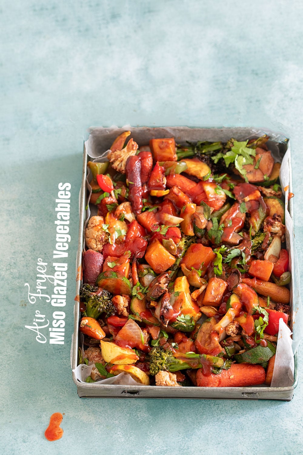 Top view of a mix of vegetables in a grey rectangle metallic tray. There is sauce drizzled on top and a drop on the outside of the tray