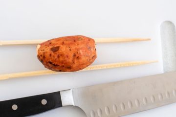 Top view of one baby sweet potato cradled by two chopsticks and sitting on a white chopping board. A large chopping knife is next to it.