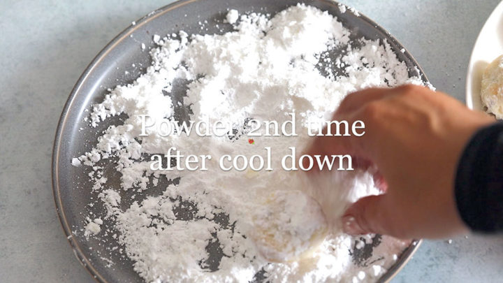 top view of the author's hand powdering the cookie on a gray plate with powdered sugar on it.