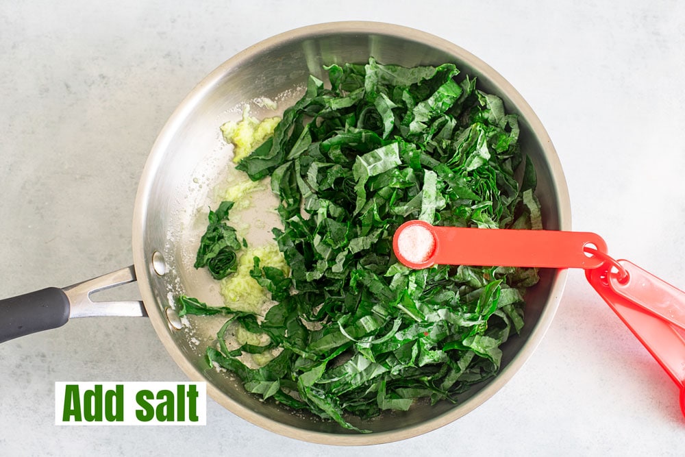 A red 1tsp measuring spoon filled with pink salt placed on top of the greens in the pan