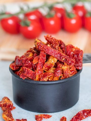 front view of sun-dried tomatoes overflowing in a black measuring cup. Tomatoes on the vine in the background