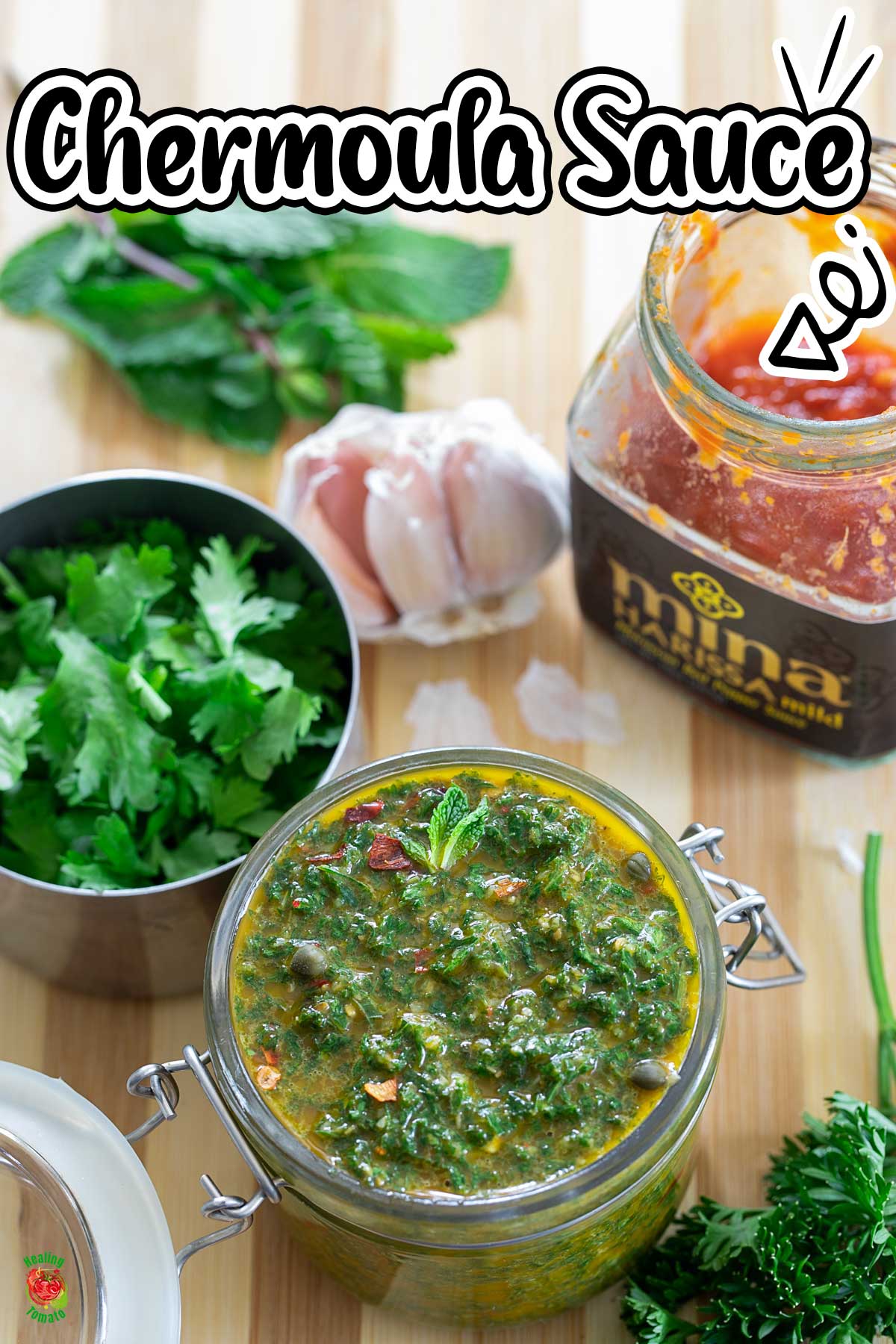 Top view of chermoula sauce in a glass jar. Jar is surrounded by fresh herbs, garlic and harissa paste.