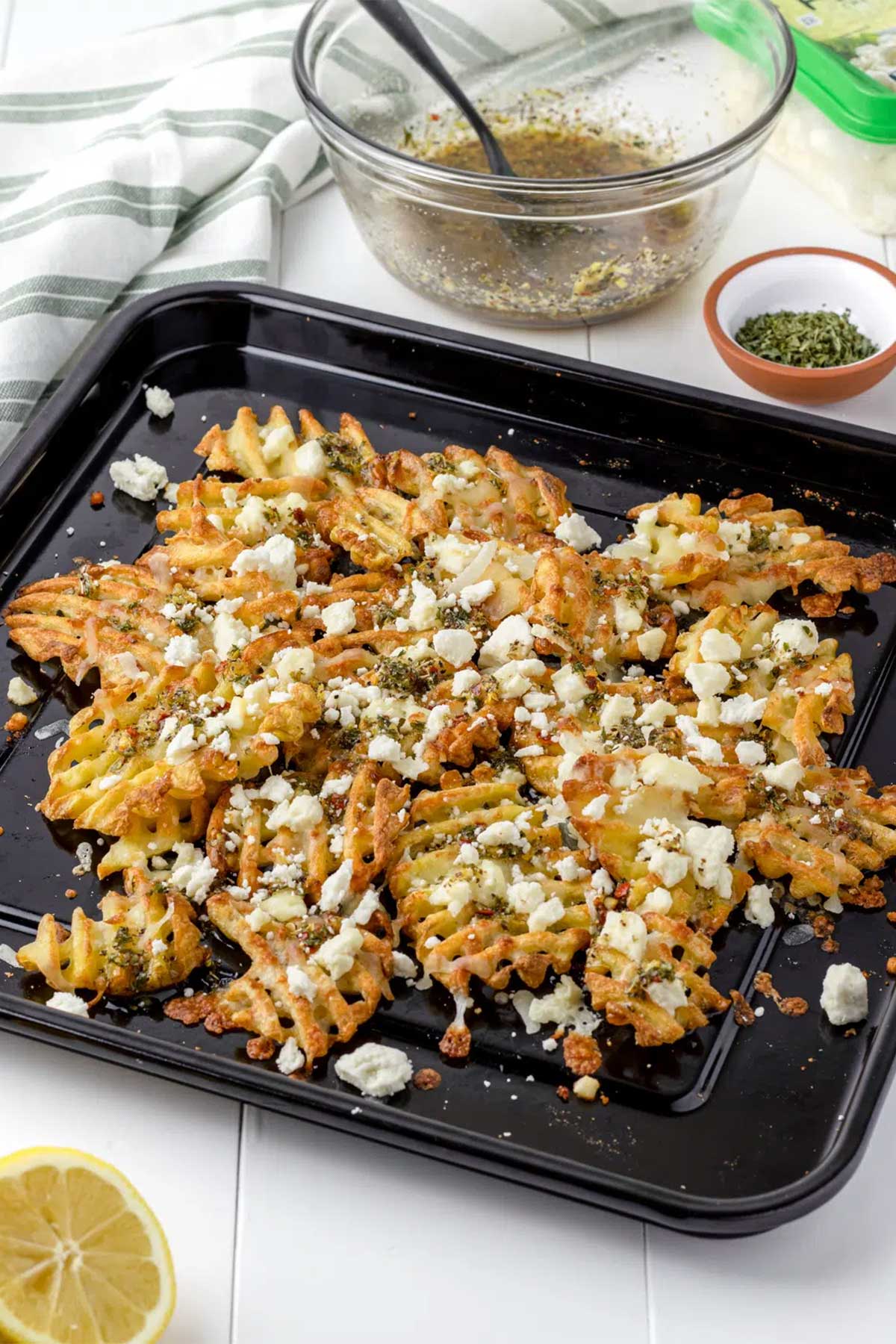 Top and angle view of waffle fries in a black sheet pan topped with feta cheese and pesto