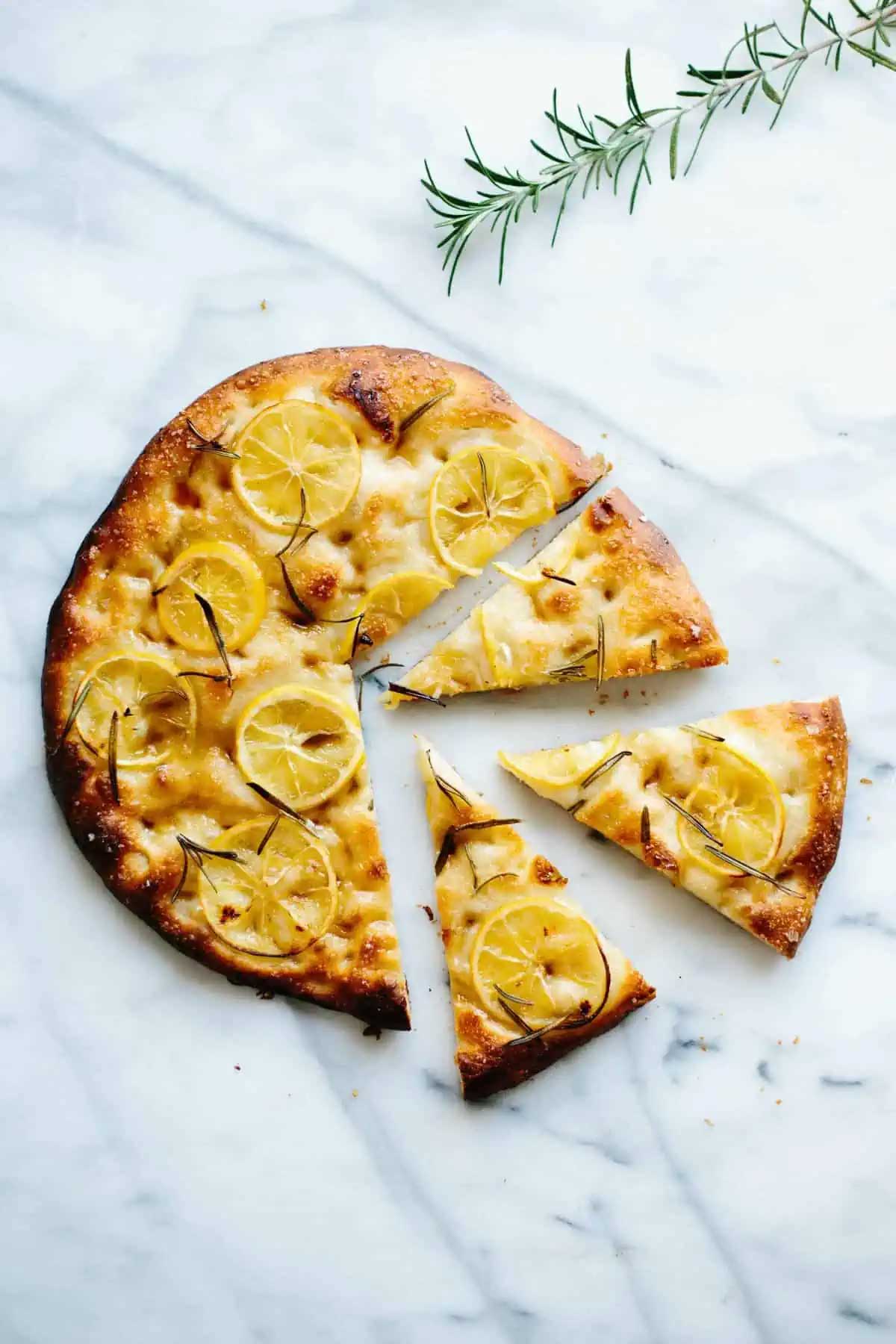Top view of a circular lemon focaccia on white marble. Half of it is cut into slices.