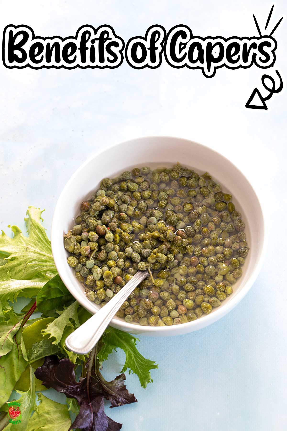 Top view of capers in a white bowl with a spoon in it. Greens on the side.