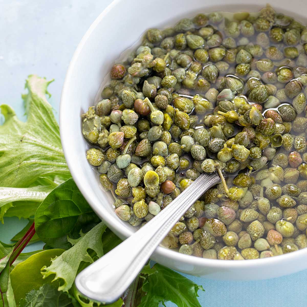 Top and closeup view of capers in a white bowl with brine.