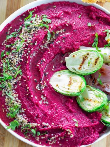 Top view of a bowl filled with vibrant magenta colored hummus. Hummus is garnished with seeds and cilantro. On the side, there are grilled veggies.