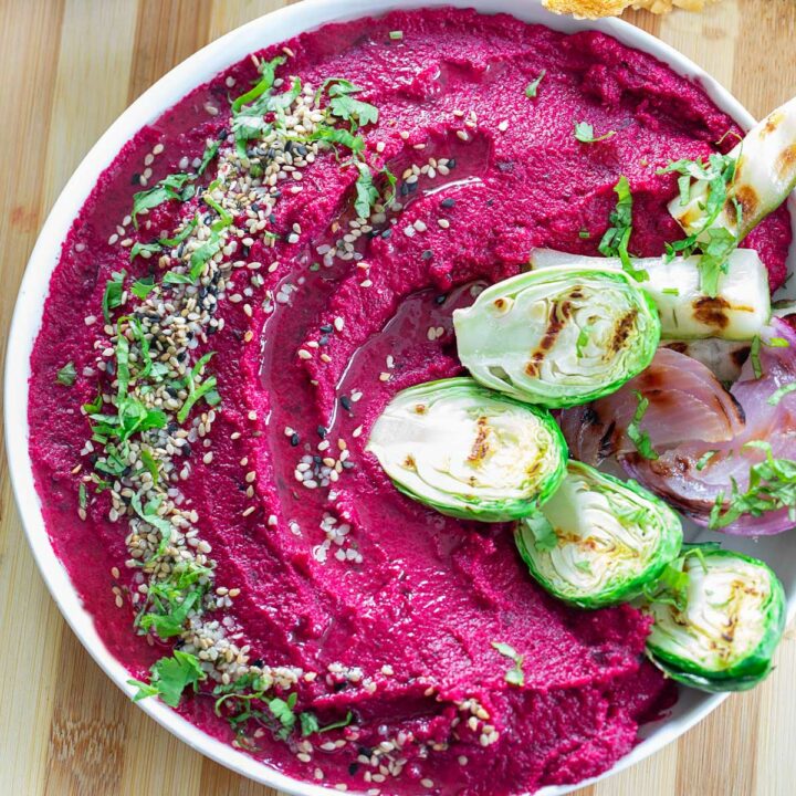 Top view of a bowl filled with vibrant magenta colored hummus. Hummus is garnished with seeds and cilantro. On the side, there are grilled veggies.