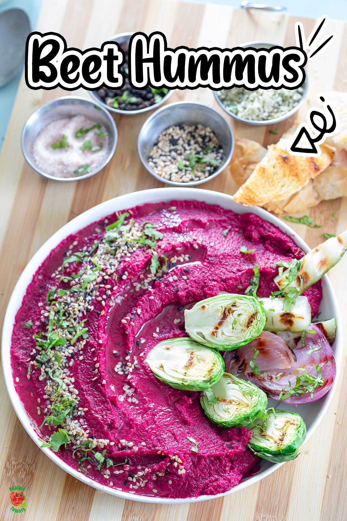 Top view of a bowl filled with vibrant magenta colored hummus. Hummus is garnished with seeds and cilantro. On the side, there are grilled veggies.