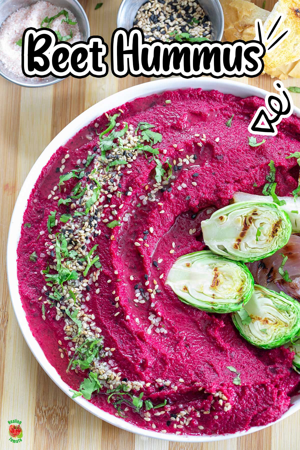 Top view of a bowl filled with vibrant magenta colored hummus. Hummus is garnished with seeds and cilantro. On the side, there are grilled veggies.