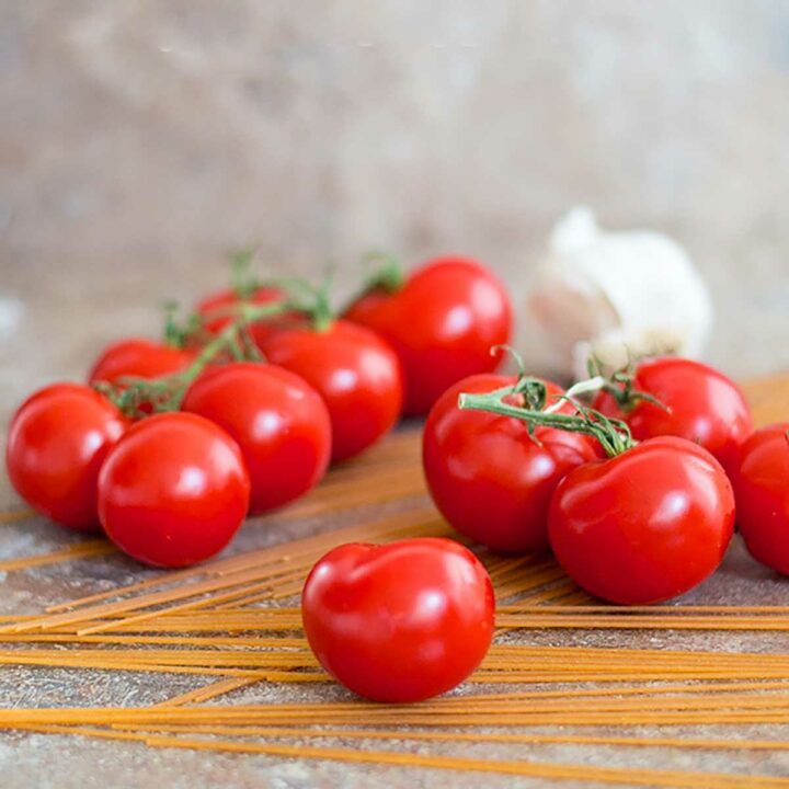 Front view of one campari tomato sitting on a few uncooked spaghetti strands. More campari tomatoes in the background