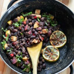 Top view of cooked Caribbean black beans in a black skillet with a wooden spoon in it and charred lime slices.
